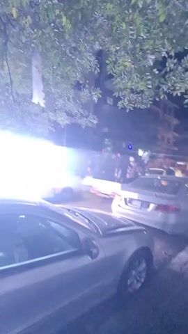 The video shows a street scene at night. There are cars parked along the street and traffic moving in the distance. In the background, there are buildings visible. There's an LED advertisement, and the horn of a car is heard. It appears to be a relatively busy street.