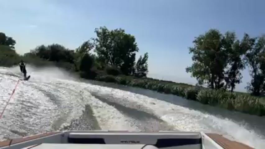 The video shows a water skier being pulled behind a boat on a river or wide channel. The skier is wearing a dark outfit and is skiing on one ski. The boat is a Nautique and is moving at a fast pace, creating a large wake. The river is lined with vegetation, including trees and tall reeds. The sky is blue with some clouds. The camera is positioned at the back of the boat, looking towards the skier. The skier falls around 0:03.