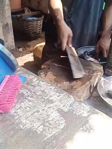 A person is chopping fish on a wooden cutting board with a cleaver. There are several fish in a metal bowl to the right of the cutting board. A pink brush is to the left of the cutting board. The scene appears to be outdoors, with a dirt floor and a wicker basket in the background.