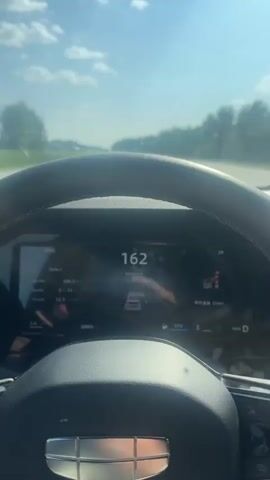 A driver's perspective video shows a car dashboard and the road ahead. The car is traveling on a highway with trees and fields lining the road under a bright, cloudy sky. The dashboard displays the speed, which fluctuates between 163 km/h and 158 km/h. Other information on the dashboard includes trip distance, fuel level, and gear selection. The driver's hands are visible on the steering wheel. The car is in motion, indicated by the changing speed and the blurry background. There are no other vehicles clearly visible in the immediate vicinity of the car.