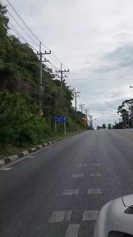 The video shows a point-of-view shot from inside a vehicle driving along a road. The road is asphalt with a white and black checkered curb on the left side. The left side of the road is bordered by a steep, lush green hillside covered in trees and vegetation. Several utility poles with wires run along the hillside. A blue sign with white text and an image is visible on the left side of the road. Further down the road, there are more utility poles and streetlights. The right side of the road curves away and appears to lead towards a distant horizon with more trees. The sky is overcast with scattered clouds. The overall impression is a scenic drive along a coastal or tropical road.