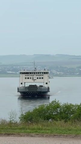 A ferry boat named ALEKSANDR ANACHKIN is traveling away from the camera on a calm body of water. The ferry is white with a dark grey hull and has several windows visible. The rear of the ferry shows stacked cargo and multiple tires along the railing. In the background, there are rolling hills and a distant town. The foreground shows green foliage and a dirt path. The sky is overcast.