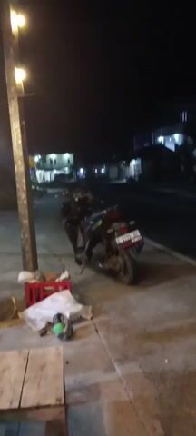 A night time shot from a sidewalk facing a street and a row of buildings. A parked motorcycle is on the sidewalk in front of a street light. There are some crates and bags on the sidewalk. The buildings appear to be houses and small businesses. The street is empty of traffic. The camera pans to the right, showing more of the street and buildings. The camera then pans left, back towards the motorcycle and sidewalk. In the foreground, a table with a glass, a pack of cigarettes and a basket of food. A red banner hangs over a stall with text that is partially obscured.