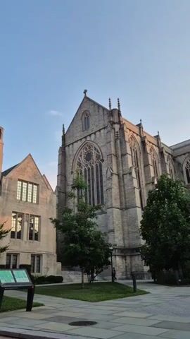 This is a vertical video shot from a street-level perspective. The scene transitions between different outdoor views of a campus or institutional setting, characterized by stone buildings with Gothic architectural elements and lush green trees.  In the first segment, two stone buildings are visible under a clear blue sky. One building on the right is larger and more ornate, with a prominent arched window and spires. A smaller building on the left features a distinctive chimney-like structure. A signpost with information is situated in the foreground, on a paved area.  The second segment shows the side of one of the stone buildings, with trees and a lamp post. The third segment shifts to a street view. A pedestrian crossing with white lines is present. A yellow diamond-shaped sign with a pedestrian symbol and a directional arrow is mounted on a pole, along with a traffic signal. Cars are driving on the asphalt road, and trees line the sidewalk. The fourth segment shows a red car turning on a street intersection, with a stone building in the background. The final segment features a wider street view with several cars, including the red car, and more trees. A basketball hoop is visible in the distance. The overall impression is a peaceful, academic or historic environment during daylight hours.