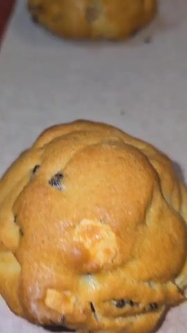 The video features a close-up view of two distinct cookies or cookie dough items on a baking sheet lined with parchment paper. Initially, a single, golden-brown baked cookie with visible chocolate chips is shown. The camera then pans to a dark red or maroon-colored raw cookie dough ball, which contains lighter-colored chunks, possibly white chocolate or nuts. After a brief focus on the raw dough, the camera moves back to highlight the baked, golden-brown cookie. The scene is set in what appears to be a kitchen, with the items resting on a baking sheet, suggesting a baking activity. There are no people visible, and no discernible text. The video has a somewhat shaky, handheld quality.