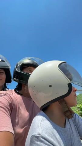 The video opens with a close-up of three people wearing helmets, appearing to be riding on a motorcycle. The person in the center, presumably the driver, looks directly at the camera. The person to their left, a woman with dark hair, is also looking towards the camera with a slight smile. The third person, in front and to the right, is mostly obscured by a white helmet. The background shows a bright blue sky. The camera then pans out to show the three individuals on the motorcycle, now with the woman on the left and the driver in the middle in clearer view. The scenery behind them consists of green foliage and trees under a clear blue sky with scattered clouds. The video then transitions to a first-person perspective of someone riding a motorcycle on a paved road. The road is lined with lush green vegetation on both sides and stretches into the distance towards rolling hills. The sky is a vibrant blue with fluffy white clouds. Shadows of trees are cast across the road, indicating sunlight. The video captures the movement of the motorcycle along the scenic route.