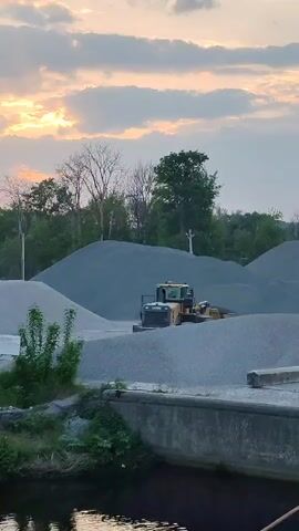 The video shows a yellow and black wheel loader operating on a large pile of gravel or crushed stone. The loader is positioned atop the gravel pile, and a person is visible standing near it, possibly observing the loader's operation. The gravel pile is part of a larger area with multiple piles of similar material, suggesting a quarry or industrial storage site. In the background, there is a line of trees and a cloudy sky with the sun setting, casting a warm glow. In the foreground, a concrete barrier or dock is present, with some vegetation growing nearby, and a dark body of water. The scene is static, with no visible camera movement or audio, capturing the loader and its surroundings in a still, scenic shot.