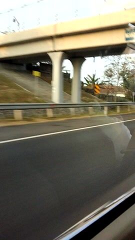 The video shows a view from a vehicle moving along a road. On the left side of the frame, an elevated structure, possibly an overpass or bridge, is visible with its concrete supports. Below this structure, there is a road or path with some vegetation and a yellow sign with a black symbol. The right side of the frame shows the road the vehicle is traveling on, which is asphalt with a white line marking its edge. The perspective suggests the camera is looking out the side window of a moving vehicle, with a slight tilt to the right. There are no people visible in the frame. The lighting suggests it is daytime.