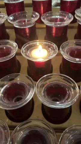 The video shows a close-up view of a row of votive candles in a church. The candles are in red glass holders with clear glass tops. One candle in the center is lit, casting a warm glow. The background is blurred, but it appears to be the interior of a church with pews and an altar visible. The camera slowly pans and zooms, focusing on the candles and the lit flame. The lighting is dim, suggesting it might be evening or a dimly lit part of the church.