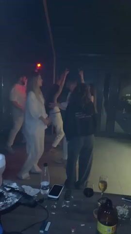 A group of young adults are dancing and enjoying themselves at a party. The room is dimly lit with red and white lights. In the foreground, there is a table with drinks, phones, and what appears to be drugs and drug paraphernalia. Some people are dancing in the background, while others are in the foreground interacting with each other. The overall atmosphere is festive and energetic.