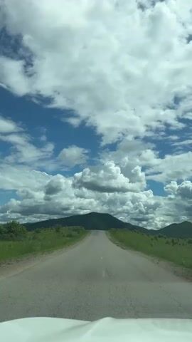 The video shows a forward-facing view from inside a vehicle on an asphalt road. The road curves slightly to the left as it leads towards a large, green hill in the distance. The sky is filled with a dramatic display of white cumulus clouds against a bright blue sky. The landscape on either side of the road is covered in green grass and some scattered bushes or low trees. The bottom edge of the frame shows the white hood of the vehicle the video is being filmed from.