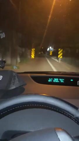 This is a first-person perspective video taken from inside a car at night. The driver's hands are on the steering wheel, which is visible in the foreground. The dashboard displays the outside temperature as 20 degrees Celsius. The car is driving down a road lined with trees and a wall on the left side. There are yellow and black striped poles visible on both sides of the road, likely marking a hazard or lane. Streetlights are visible in the distance, illuminating the road ahead. The overall impression is of a quiet drive on a suburban or rural road at night.