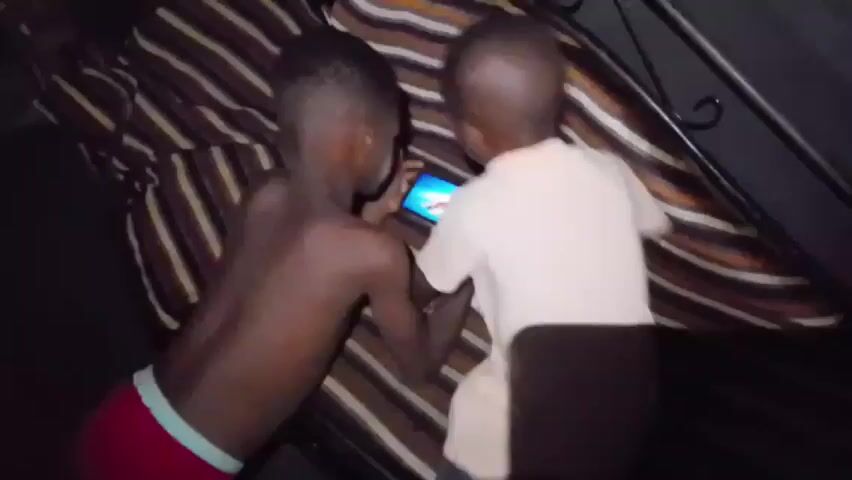 Two young, dark-skinned boys are in a dimly lit area. One boy is lying on his back on a striped cushion, wearing only red underwear. The other boy, wearing a white shirt and navy shorts, is standing near him. The boy on the cushion holds a smartphone in his hands and looks directly at the camera. The other boy watches the phone with a curious expression. The environment appears to be indoors, possibly a tent or under a bed frame, as evidenced by the dark surroundings and the presence of metal bars in the background.