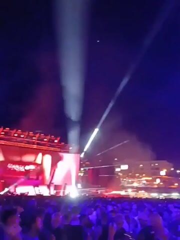 The video shows a large crowd of people at an outdoor music event at night. The crowd is dense, and the view is from within the crowd, looking towards the stage. Bright white and red spotlights illuminate the stage area and the crowd. A building with lights on is visible in the background, behind the stage. The atmosphere appears lively and energetic.