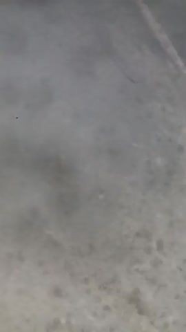 The video shows a close-up, blurry view of a gray, textured surface, possibly concrete or stone. Wisps of white smoke or fog drift across the bottom of the frame, obscuring portions of the surface. The lighting is dim, and the overall impression is one of muted tones and limited detail.