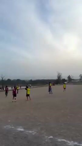 The video shows a sports field on a cloudy day. Several young people are playing a sport, possibly soccer, on a dirt field. Some are wearing yellow jerseys, and others are wearing red and black striped jerseys. A group of people are sitting on a low wall along the edge of the field, watching the game. There are trees and buildings in the background, and a distant view of a city or town. The sky is overcast with the sun trying to peek through the clouds. The scene appears to be a casual, local sporting event.