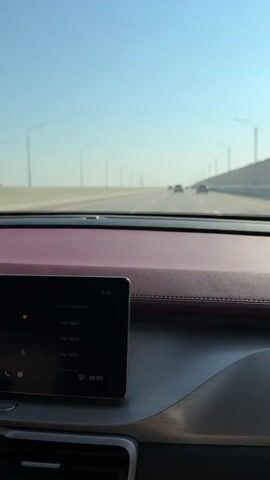 The video shows a view from inside a car, looking out at a highway. The dashboard is visible in the foreground, with a touchscreen display showing radio stations and other information. A hand is seen interacting with the touchscreen. The road ahead is clear with a few other cars in the distance. The sky is bright blue. The car appears to be moving on a multi-lane highway with streetlights lining the sides.