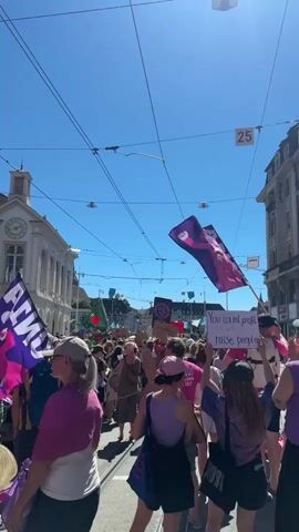 A group of people are marching down a street. They are holding flags and signs. One sign reads "You count people, raise people." The flags are pink and purple. The people are wearing a variety of clothing. The sky is blue and clear. There are buildings on either side of the street. There are also power lines overhead. The street appears to be paved with cobblestones or similar material. Some of the people are holding up their hands. The video is shaky, suggesting it was filmed by someone walking or moving.