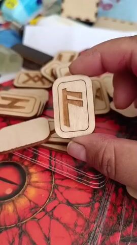 A hand holds up a wooden rune with the symbol F carved into it. In the background, more runes are visible, along with other objects. The rune is rectangular with rounded corners and a border. The symbol F is carved into the wood, appearing as a vertical line with two horizontal lines extending from the right side. The background is blurred, but it appears to be a surface with a red and black pattern and some other items.