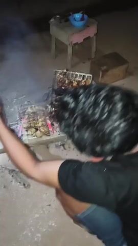 The video shows a person grilling food over hot coals. The food appears to be chicken pieces on a metal grill. Some of the chicken is well-cooked and charred, while other pieces are still cooking. There are also some vegetables, possibly potatoes, on another grill. The scene is outdoors, and the lighting suggests it is nighttime. There is smoke rising from the coals. The person is holding the grill with a wooden handle.
