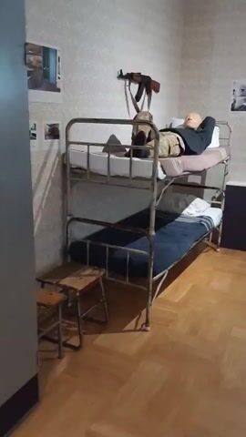 This media file shows a dimly lit room with bunk beds. The beds are made of aged, possibly rusty metal, with worn mattresses. The upper bunk is occupied by a mannequin dressed in military-style camouflage clothing, with a rifle mounted on the wall above it. The mannequin has its head resting on a pillow, appearing to be sleeping or resting. A backpack is placed on the upper bunk next to the mannequin. The lower bunk is made with a dark blue blanket and a white pillow. Wooden stools are positioned in front of the bunk beds. The room has patterned wallpaper and a wooden floor. The overall scene appears to be a museum exhibit or a recreated living space, possibly depicting a barracks or a soldier's quarters, given the military elements. The lighting suggests it is indoors, and there are no clear indications of the time of day or weather. The location context is Viimsi, Estonia, which could imply a historical or military museum exhibit related to Estonia's past. There is no visible text in the media file.