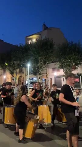 This video captures a lively street performance in Arenys de Mar, Spain, as dusk settles. A group of young people, dressed in black t-shirts adorned with a green and red emblem, are energetically playing drums. The drums, mostly large, yellow, barrel-shaped instruments, are positioned in a semi-circle, with the performers taking turns striking them with drumsticks. The expressions on the faces of the drummers suggest focus and enjoyment, contributing to an enthusiastic atmosphere.

The scene unfolds on a street lined with trees, their leaves creating a dappled effect against the twilight sky. Buildings with balconies and illuminated windows are visible in the background, hinting at a residential or commercial area. Streetlights cast a warm glow, adding to the ambiance of the evening. The sky is a deep blue, indicating that it is either early evening or late twilight.

A few onlookers are present, observing the performance. The presence of a street sign with what appears to be Spanish text, and the architectural style of the buildings, confirm the location in Spain. The overall mood is festive and engaging, with the rhythmic drumming creating a dynamic and memorable street spectacle. No other text is clearly legible in the video beyond the emblem on the t-shirts.