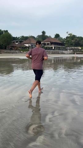 The video shows a man running barefoot on a wet beach. The man is wearing a dark shirt and shorts. He is running away from the camera. The beach is wide and flat, and the water is shallow. There are buildings and trees in the background. The sky is overcast. There is also someone else on the beach further away. The man's reflection is visible in the water.