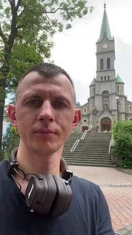 A man with short hair and headphones around his neck stands in front of a church with a tall steeple. He is facing the camera, looking directly at it. The church has large stone steps leading up to its entrance. Lush green trees surround the church and the man. The sky is cloudy. The man is wearing a dark t-shirt. The headphones are brown and have the "Marshall" logo visible. The scene appears to be outdoors during the daytime.