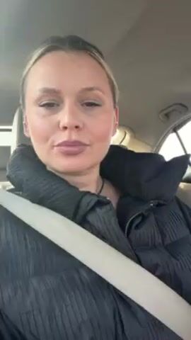 A woman is recording a video from the passenger seat of a car, wearing a black puffer jacket. She looks directly at the camera and smiles. The driver is a man wearing sunglasses and a black and white tracksuit, looking ahead. There is a dog in the back seat. The car is driving on a road with snow-covered ground visible through the windows. There is music playing in the background.
