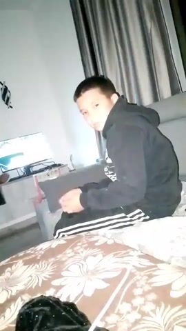 A young boy wearing a black hoodie with white stripes sits on a couch. He is holding a white object in his hands. Two televisions are visible in the background. The room appears to be a living room. The lighting is dim.