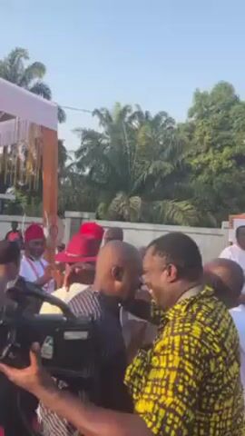 The video shows a gathering of people outdoors on a sunny day. Some of the men are wearing traditional attire, including red hats. Some individuals greet each other with hugs.  A man with a camera is filming. A white tent or structure with drapes is visible in the background. There are trees and a building in the background. The overall atmosphere appears celebratory or social.