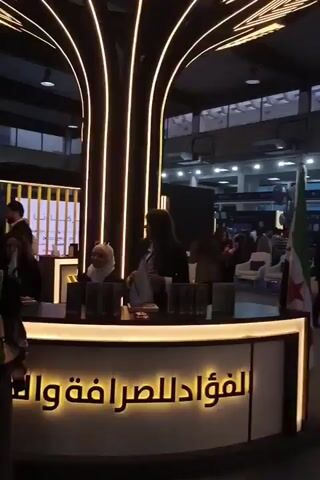The video depicts an indoor exhibition or trade show environment. The camera moves through a crowd of people, most of whom are dressed in business attire, walking and interacting. Booths are visible on either side of the pathway, with signage in Arabic and displays promoting various companies or organizations.  The lighting is bright, and the ceiling is high with industrial-style ventilation. There is a flag of Syria is displayed at one booth.