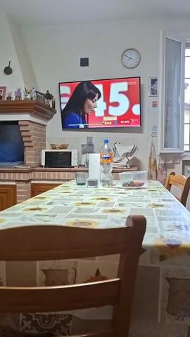 A person is sitting at a wooden dining table covered with a sunflower-patterned tablecloth. In the background, a flat-screen television mounted on the wall shows a news broadcast with a woman speaking. Below the television, a brick-built fireplace is visible, topped with a mantelpiece adorned with framed photos and decorative items. To the right of the fireplace, a white microwave oven sits on a countertop. On the table, a plastic bottle of Fanta, a glass, and a bowl of fruit are present. A window with white curtains is visible on the left side of the frame.