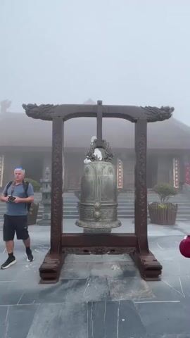 The video begins with a close-up of a large, ornate bronze bell mounted on a wooden frame. The bell is detailed with intricate carvings and appears to be outdoors, with a misty, overcast sky in the background. In the foreground, a man in a blue shirt and shorts walks towards the left side of the frame, carrying a backpack. To the right of the bell, a person in a maroon jacket and a wide-brimmed hat is standing with their hands clasped in front of them, facing the bell. As the video progresses, the camera pans slightly to the right, revealing more of the surrounding environment. A traditional East Asian-style building with a curved roof and decorative eaves is visible in the background, partially obscured by mist. Several potted plants are placed around the area, and there are steps leading up to the building. Other people are scattered throughout the scene, some standing, some sitting, and some walking. The ground is paved with large, grey stone tiles. The overall atmosphere is serene and slightly somber due to the misty conditions. The video captures a moment of quiet contemplation and tourism at a scenic and culturally significant location.