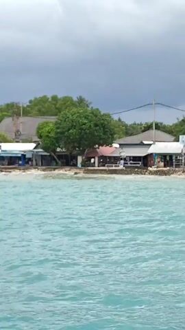 The video shows a seascape of aqua-blue waters with several boats bobbing near a shore. The beach is lined with buildings and vegetation under a cloudy sky. Various boats, including small motorboats and larger vessels, are seen scattered across the water. The shoreline features structures that appear to be a mix of commercial buildings and traditional thatched-roof huts, nestled among lush green trees. The overall scene suggests a coastal tourist destination, possibly an island or a resort area, with a calm, albeit overcast, weather condition.
