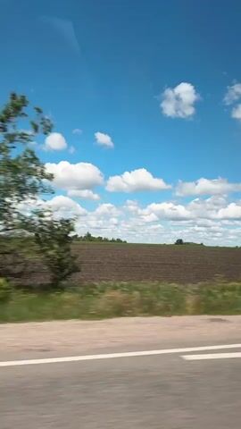 The video shows a view from a moving vehicle, likely a car or bus, driving along a road. The road is gray asphalt with white lane markings. Adjacent to the road is a strip of green vegetation, followed by a large, plowed field of dark brown soil. The sky is a bright blue, scattered with numerous fluffy white clouds. A vapor trail is visible in the sky, likely from an airplane. The camera is positioned inside the vehicle, showing a clear view of the landscape passing by. The overall scene is rural, presenting an open, agricultural landscape.