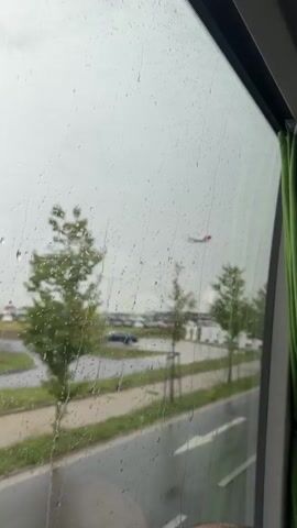 The video shows a view through a wet window, with raindrops obscuring the scene outside. In the distance, an airplane is visible in the sky, seemingly ascending or descending. The landscape outside includes trees, a parking lot with vehicles, lampposts, and some buildings. The weather appears to be overcast and rainy. The perspective suggests the viewer is inside a vehicle or building, looking out.