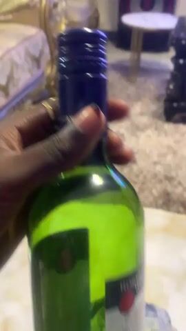 A dark-skinned hand holds a green wine bottle labeled "ROBERTSON WINERY CHAPEL SWEET RED" containing red wine, then places it upright on a dark brown wooden side table. The hand then picks up a clear stemmed wine glass containing a small amount of red wine. The scene is indoors, featuring a light-colored tiled floor, a brown shaggy rug, and an ornate gold-trimmed sofa.