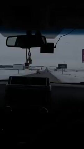 The video is shot from the interior of a vehicle driving down a road. Snow-covered fields can be seen on either side of the road. The sky is overcast. The dashboard, rearview mirror, and a dashcam are visible in the foreground, creating a darkly lit interior view. Other vehicles can be seen driving on the road ahead. Two large billboards are visible along the roadside.