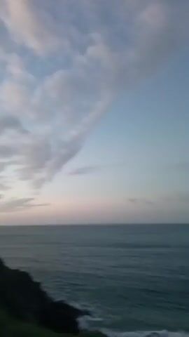 The video shows a scenic view of an ocean and a sandy beach at dusk. The camera initially focuses on the horizon, revealing a calm ocean under a sky with soft, pastel-colored clouds. It then pans to the right to show a bay, revealing a small island and a coastline of hilly terrain covered in vegetation. The camera movement pauses, bringing into view a stretch of sandy beach with gentle waves lapping at the shore. Finally, a woman with dark hair and fair skin appears on the right side of the frame, looking out at the view. She is standing slightly apart from the camera, and her expression appears thoughtful as she observes the landscape. The general tone of the video is peaceful and contemplative.