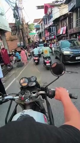 A first-person perspective video shows a person riding a motorcycle through a crowded street lined with shops. Several other motorcycles and scooters are also present, as well as a few cars. People are walking on the sidewalks and in the street. The buildings have signs for shops and a hotel. The street is paved and there are utility wires overhead. The time is daytime.