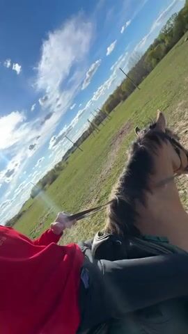 A person is riding a horse across an open, grassy field under a bright blue sky with scattered white clouds. The camera is positioned from the rider's perspective, looking forward over the horse's neck. The rider wears a red sweatshirt with "5 DES" visible text and black riding pants, holding the reins. The horse, a light brown color with a darker mane, is fitted with a bridle and saddle and appears to be walking or trotting gently. In the background, there are several wooden poles stretching across the field, and trees line the distant horizon. The sun is shining brightly, causing some lens flare. The setting appears to be a rural or equestrian area, specifically located in Капцёўскі сельскі Савет, Belarus.