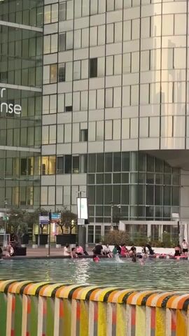 This video shows a wide shot of a public plaza in front of a modern, glass-fronted building. In the foreground, a colorful, striped barrier lines the edge of a shallow body of water, possibly a fountain or reflecting pool. A group of people are seated along the edge of the water, with some dipping their feet in. The group appears to be diverse in age and is engaged in casual conversation and relaxation. Some individuals are wearing brightly colored clothing.

Behind the water feature, the main subject is a large, multi-story building with a distinctive curved glass facade. The windows reflect the sky and surrounding environment, giving the building a sleek, contemporary appearance. Below the main glass structure, there is a section with a white facade, featuring a restaurant or cafe with outdoor seating visible through the windows. A sign above this section reads "ECLIPSE" in white lettering. Another sign below that reads "KOZEDO."

The lighting suggests it is late afternoon or early evening, with warm light emanating from the restaurant and illuminating some of the people. The overall atmosphere appears relaxed and pleasant, with people enjoying a leisurely time outdoors in an urban setting. The presence of the modern architecture and the public gathering space suggests a vibrant city center. There are a few trees and streetlights visible in the background, further establishing the urban environment.