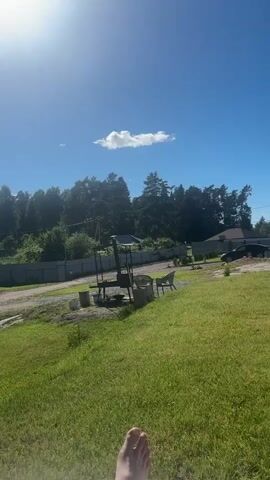 This is a vertical video captured on a sunny day in Khvoynoye, Russia. The scene appears to be a rural or suburban residential area with a large grassy yard in the foreground. In the distance, there's a line of trees, a fence, some houses, and a road.

The video shows a foot resting on a chair in the foreground, suggesting someone is relaxing outdoors. In the middle ground, there's a metal barbecue grill with some firewood and chairs next to it, indicating outdoor cooking or leisure activities. Further back, a black car is parked. On the right side of the video, a modern white building with a wooden deck is visible. An orange stand-up paddleboard and a paddle are leaning against the wall of this building, implying recreational water activities might be planned or have recently taken place.

The sky is a clear, bright blue with a single small white cloud. The sun is high and bright, casting shadows and indicating it's likely midday. The overall mood is peaceful and leisurely, suggesting a summer day spent enjoying the outdoors. There is no visible text in the video.