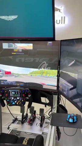 This media file displays a high-end racing simulator setup, likely in a home or dedicated sim racing room, given the presence of a keyboard and multiple monitors. The primary focus is on the simulation equipment itself, not on any individuals.

The scene is dominated by a sophisticated racing wheel with an integrated digital display and numerous buttons, suggesting it's designed for serious sim racing. Attached to the wheel are three pedals: clutch, brake, and accelerator. The entire setup is mounted on a robust aluminum rig, indicating a commitment to realism and stability.

The monitors are a key feature. A large, curved ultra-wide monitor displays a racing game, showing a track with green and blue curbs, grassy verges, and trees in the background. The in-game interface shows speed and other telemetry data. Above this, a second monitor displays the Aston Martin logo. To the left of the main display, another monitor shows what appears to be a different view or menu from the game, possibly a side mirror or data display. A keyboard is positioned to the left of the steering wheel.

The lighting suggests an indoor environment, possibly with overhead room lighting. There are no clear indicators of the time of day or weather outside. The overall impression is one of a dedicated and well-equipped space for virtual motorsport.

Visible text includes the "Aston Martin" logo on the top monitor, and within the racing game on the main monitor, there are various in-game graphics and text elements indicating speed, lap information, and potentially driver names or car settings, though these are not clearly legible. A "Red Bull" logo is also visible in the background on a wall-mounted sign.