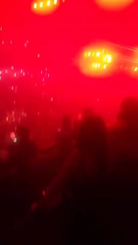 The video shows a concert or event venue with a large crowd. The video is shot from an elevated perspective, possibly a balcony or platform, behind a group of people. The lighting alternates between bright white and intense red, creating a dramatic effect. The crowd is dense, and many people are holding up phones, seemingly recording the event. The stage, visible in the distance, is lit with several bright lights and appears to have an active performance ongoing. The atmosphere is filled with smoke or fog, enhancing the visual effects.