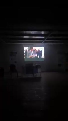A video is being projected in a dimly lit room. The projection shows a man in a suit signing a document, and other people are standing nearby. In front of the projector is a table with an Erasmus+ banner. There are chairs and posters along the walls of the room. The ceiling is visible with what appears to be piping or wiring.