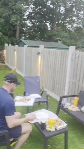 The video shows an outdoor scene on a lawn area with artificial turf. In the foreground, there is a small black coffee table with a white plate, a container of food, a drink, and two bags of chips on it. A person is sitting in a dark blue chair to the left of the table, appearing to be preparing food or a plate. To the right of the table, there is a dark bench with several bags of chips and a drink on it. Behind the bench and table is a wooden fence. Two dark blue chairs are placed near the fence. The scene appears to be a casual outdoor gathering or meal. The video has some motion blur, particularly when a person is shown moving.