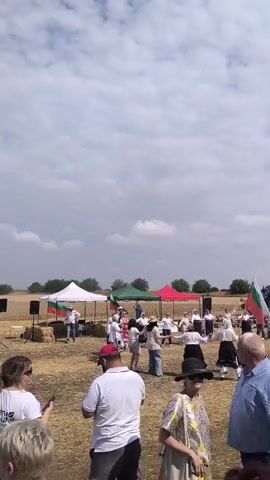 This media file shows an outdoor event in Koshava, Bulgaria. The scene appears to be a field, likely a farm, with harvested stubble. In the background, a large green and white combine harvester is parked. Several people are gathered in the field, dressed in casual attire. Some are standing around, while others appear to be participating in activities.

In the foreground, a group of people are visible. One woman in sunglasses is looking at her phone. Another woman is wearing a white top and holding a phone. A man in a red baseball cap and white polo shirt is facing away from the camera. Another man, wearing a patterned top and a black hat, is also facing away.

Further back, closer to the combine harvester, there are several tents set up. These tents are white, green, and red. People are seen standing near these tents, and some appear to be engaged in conversation or watching something. There is also a small group of children in white shirts and shorts, some of whom are kicking a ball. In the distance, there are trees and a few small buildings.

The event seems to be taking place during the day, with a bright sky filled with scattered clouds. The weather appears to be warm and sunny. The overall atmosphere seems relaxed and festive, suggesting a community gathering or a local festival. The combine harvester and the field setting indicate a possible agricultural theme to the event.

There is some visible text on the side of the combine harvester: "CLAAS."