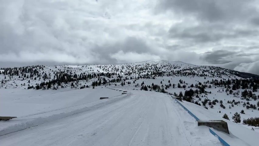 The video shows a snowy, wintery landscape under a cloudy sky. The foreground features a snow-covered track or road with visible tire marks, flanked by concrete structures painted with a blue stripe. Beyond the track, a snow-covered field slopes upwards towards a hill or mountain densely populated with evergreen trees. The trees are also dusted with snow. In the distance, a line of mountains is visible under a mix of clouds and patches of blue sky. The scene is quiet and still, depicting a remote, cold environment.